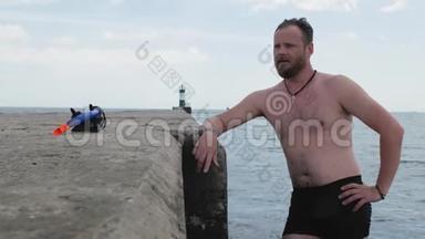 一位留着胡须的男游泳运动员站在一个混凝<strong>土</strong>码头上，在灯塔的背景下在海里游泳。 4k. 4k<strong>视频</strong>