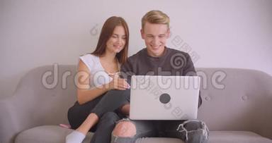 一对年轻可爱的白种人夫妇用笔记本电脑一<strong>起坐</strong>在公寓里的沙发上的特写照片