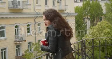 穿着皮夹克的漂亮黑发女孩，从阳台上观看绿色城市<strong>景色</strong>，喝着热茶，是个<strong>梦幻</strong>般的女孩