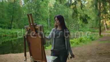 美丽的女艺术家在池塘附近的公园里画一幅风景画