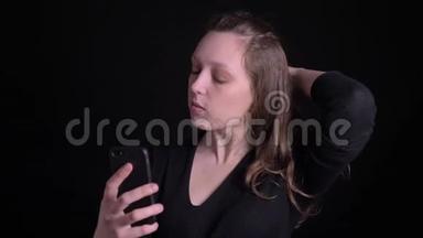 年轻卷发女孩在黑色背景下愉快地使用智能手机制作自拍照片的肖像。