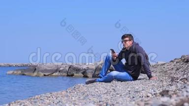 人类旅行者坐在海石海滩上，微笑和在智能手机上输入信息。