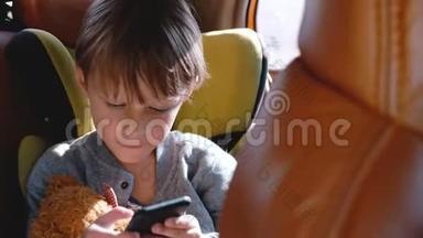 快乐兴奋的4-6岁白种人男孩微笑，使用智能手机坐在汽车儿童安全座椅与玩具。