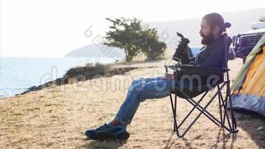 年轻人坐在湖岸露营地的椅子上，<strong>喝</strong>着<strong>热水</strong>瓶