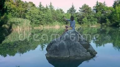 一个年轻人坐在湖中央的石头上，自拍。 使用智能手机进行摄影。 的概念