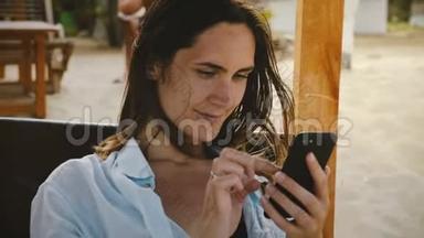 美丽的电影镜头，快乐的年轻女子使用智能<strong>手机电</strong>子商务应用程序微笑，放松休息在海滩椅子。