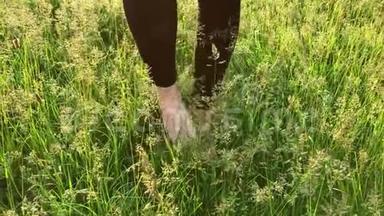 在阳光明媚的日子里，一个穿着黑色裤子站在绿草上揉搓腿的女孩光着脚的特写镜头