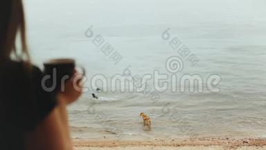 独自一人的女人早上在阳台上喝咖啡或茶，有蓝色的海景，狗在海<strong>水中玩耍</strong>，快速的视频