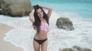 慢动作，年轻的微笑模特穿着泳装在海滩上摆着白色的沙子和大浪，美丽的岩石