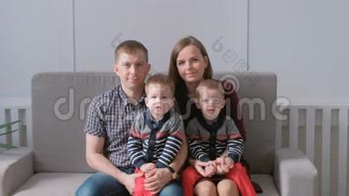 家庭视频肖像妈妈，爸爸和两个双胞胎兄弟蹒跚学步。 他们坐在沙发上看着摄像机。