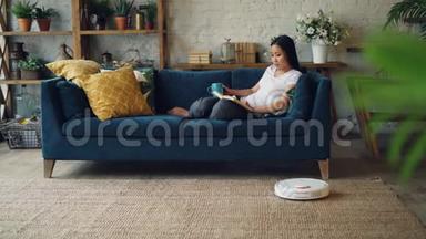 放松的亚洲女人正在看书，在家里坐在沙发上翻书页，机器人胡佛正在打扫地板