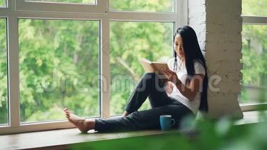 迷人的亚洲女学生正在阅读书籍，微笑着坐在现代公寓的窗台上。 爱好，青年