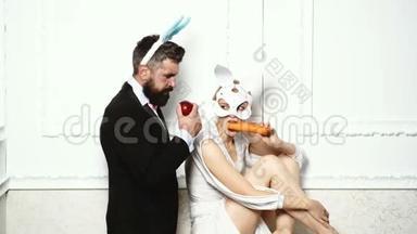 一个长胡子的男人穿着西装，吃着一个苹果，站在一个女人旁边，她戴着兔子面具，吃着胡萝卜