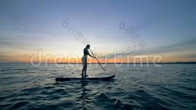 日落水景，一位苗条的女士站在木板上划水。 自由概念。