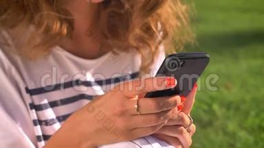 年轻的白种人黑发女人坐在草地上的公园里，用智能手机，滚动，打字，背景是大学