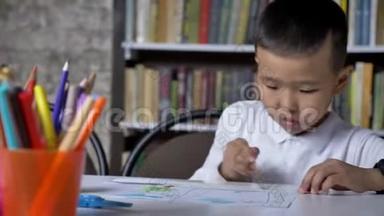 年轻的亚洲小男孩在纸上画画，坐在桌子后面，孩子做作业，书架背景