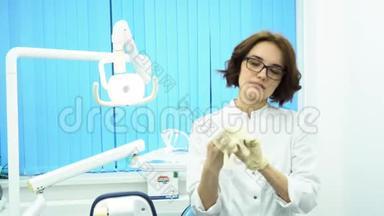 年轻的女医生戴着眼镜和一件白色的外套，在牙科诊所的戴上白色的医疗手套。 严格的