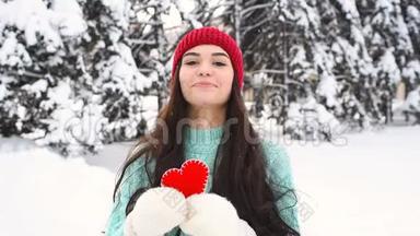 年轻漂亮的女孩穿着一件温暖的蓝色毛衣，戴着手套，背景是白雪覆盖的树木，抱着一颗红色的心，