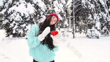 年轻漂亮的女孩穿着一件温<strong>暖</strong>的蓝色毛衣，戴着手套，<strong>背景</strong>是白雪覆盖的树木，抱着一颗红色的<strong>心</strong>，