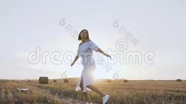 一个年轻可爱的女人在日落时快乐地穿过一片金色的田野，带着草捆。 快乐的女孩享受和奔跑