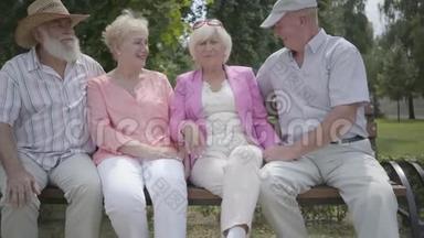 两对可爱的成熟夫妇在夏天公园里坐在长凳上聊天和微笑。 老年夫妇双人约会