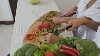 厨房桌子上砧板的俯视图。女人的手把青椒和红椒切成两半作为蔬菜