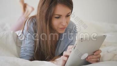 穿着蓝色衬衫，棕色头发的女人躺在床上，铺着白色床单，上网冲浪