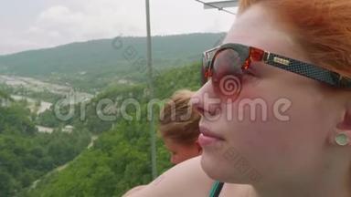 戴着墨镜的红发白种人女人看着来自高山悬索桥的蹦极。 全景