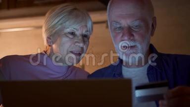 一对老夫妇晚上在家厨房。 退休家庭用笔记本电脑上网购物