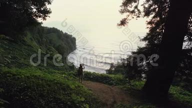 旅游女孩在智能手机上拍照，站在悬崖山上，日落时，山林中的海浪