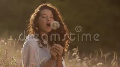 美丽的年轻女子在夏日夕阳下的麦田里吹蒲公英。 美丽与夏日概念