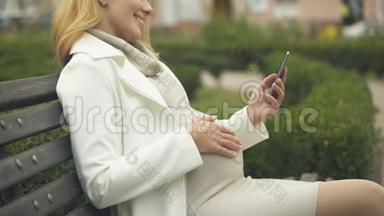 快乐的<strong>孕妇</strong>滚动智能手机<strong>照片</strong>，在长凳上的公园放松