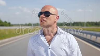 一个戴太阳镜的秃头男人在路上唱歌和散步的肖像。 中弹。