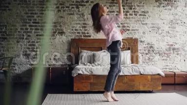 有趣的女孩正在跳舞和唱歌，拿着吹风机在卧室的地毯上玩得很开心，靠近双人床。 活跃的年轻妇女