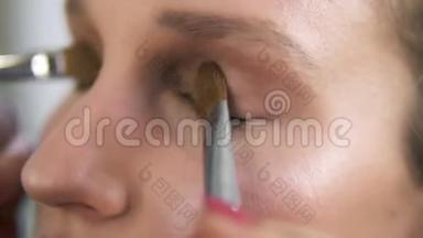 化妆师用两把刷子把浅棕色的阴影涂在眼睑上，合上一个年轻的白种人女人