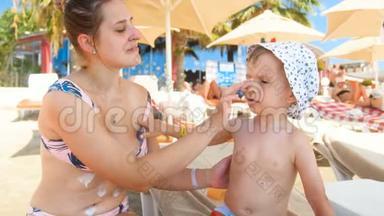 4k镜头，微笑的母亲保护她的孩子免受阳光紫外线的照射，并在海滩上涂上<strong>防晒乳</strong>液