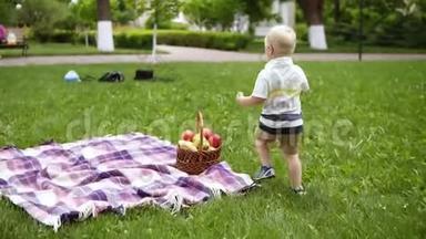 一个金发男孩在绿草上奔跑的慢动作。 从野餐篮子里拿出一个红苹果送给他的爱人
