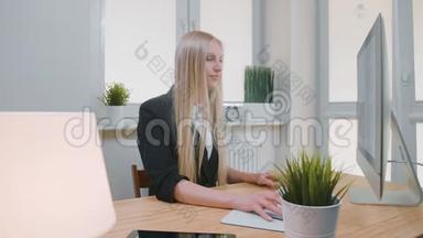 微笑的女人在办公室里用电脑工作。 年轻漂亮的金发女郎坐在工作场所，拿着电脑