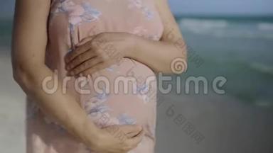 年轻的孕妇站在<strong>蓝色</strong>海洋附近的海滩上呼吸，靠近手和腹部。 暑假假期