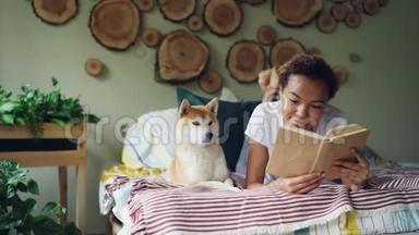 微笑的非裔美国学生可爱的女孩正在家里的床上看书，而她的宠物狗躺在她附近。 霍比特人