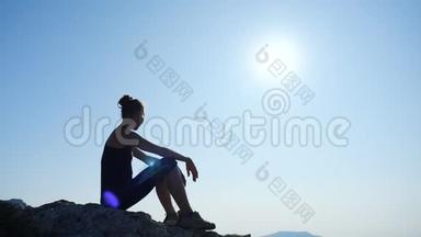 年轻受启发的女人坐在海面上<strong>一座山</strong>的顶部，对抗美丽的蓝天。 快乐的剪影