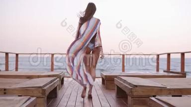 华丽的背面慢动作镜头，一个穿着泳衣的白种人模特和夏季开衫走在木地板上
