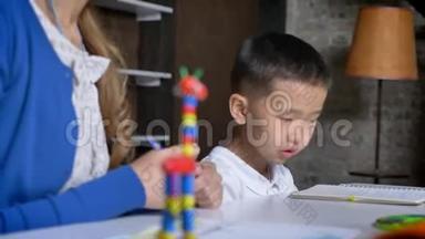 亚洲小男孩从地板上拿着落下来的笔，和母亲一起学习，坐在桌子、书架和砖墙后面