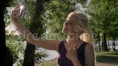 在<strong>河边公园</strong>，一位年轻的金发女子用手机自拍、竖起大拇指并微笑的肖像