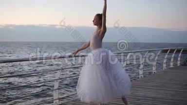 穿着白色芭蕾舞裙的优雅芭蕾舞女在靠近大海的横杆前伸展。 美丽的年轻芭蕾舞演员