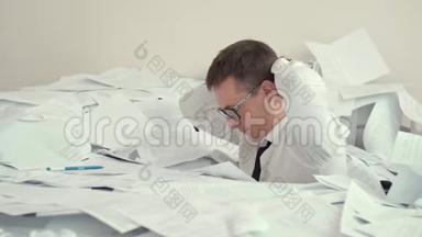 一个戴眼镜的疲惫的年轻人坐在一堆文件和签名文件里。 因为疲劳，一个男人看起来很有希望