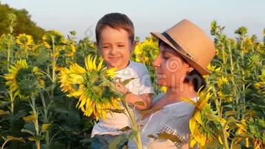 快乐的妈妈和宝宝在夕阳下看着田野里一朵鲜艳的黄色向日葵花。 一个小男孩坐在他的