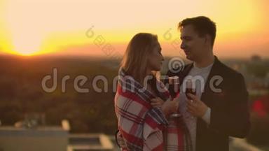 男人和年轻漂亮的女人坐在沙发上，拿着酒杯，在日落时亲吻屋顶露台。