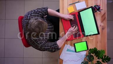 一位年轻的高加索商人在办公室里用绿色屏幕的平板电脑在笔记本电脑上打字的特写镜头
