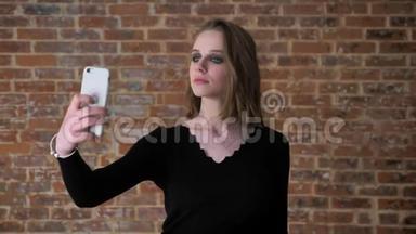 有着烟熏眼睛的年轻女孩在她的智能手机上制作seifie，触摸头发，交流理念，砖背景